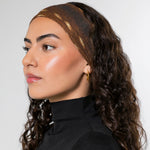 A woman with long, curly dark brown hair is shown in profile wearing a textured brown headband with gold  accents and a black turtleneck top against a plain light gray background.