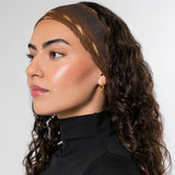 A woman with long, curly dark brown hair is shown in profile wearing a textured brown headband with gold  accents and a black turtleneck top against a plain light gray background.