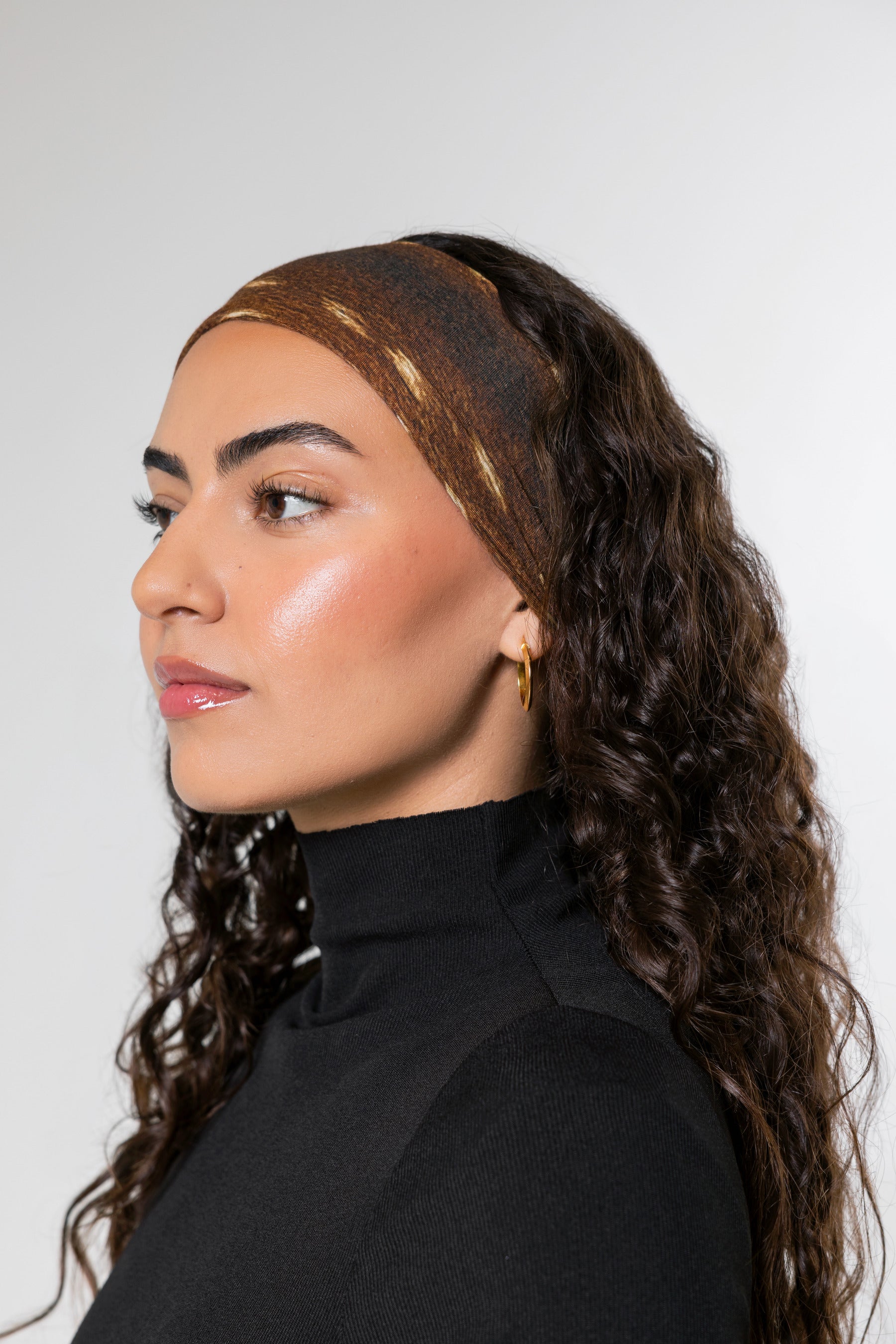 A woman with long, curly dark brown hair is shown in profile wearing a textured brown headband with gold  accents and a black turtleneck top against a plain light gray background.