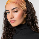 A woman with long, curly dark brown hair wearing a wide, soft fabric headband in warm gradient tones of orange, yellow, and peach, paired with a black turtleneck top against a light gray studio background.