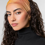 A woman with long, curly dark brown hair wearing a wide, soft fabric headband in warm gradient tones of orange, yellow, and peach, paired with a black turtleneck top against a light gray studio background.