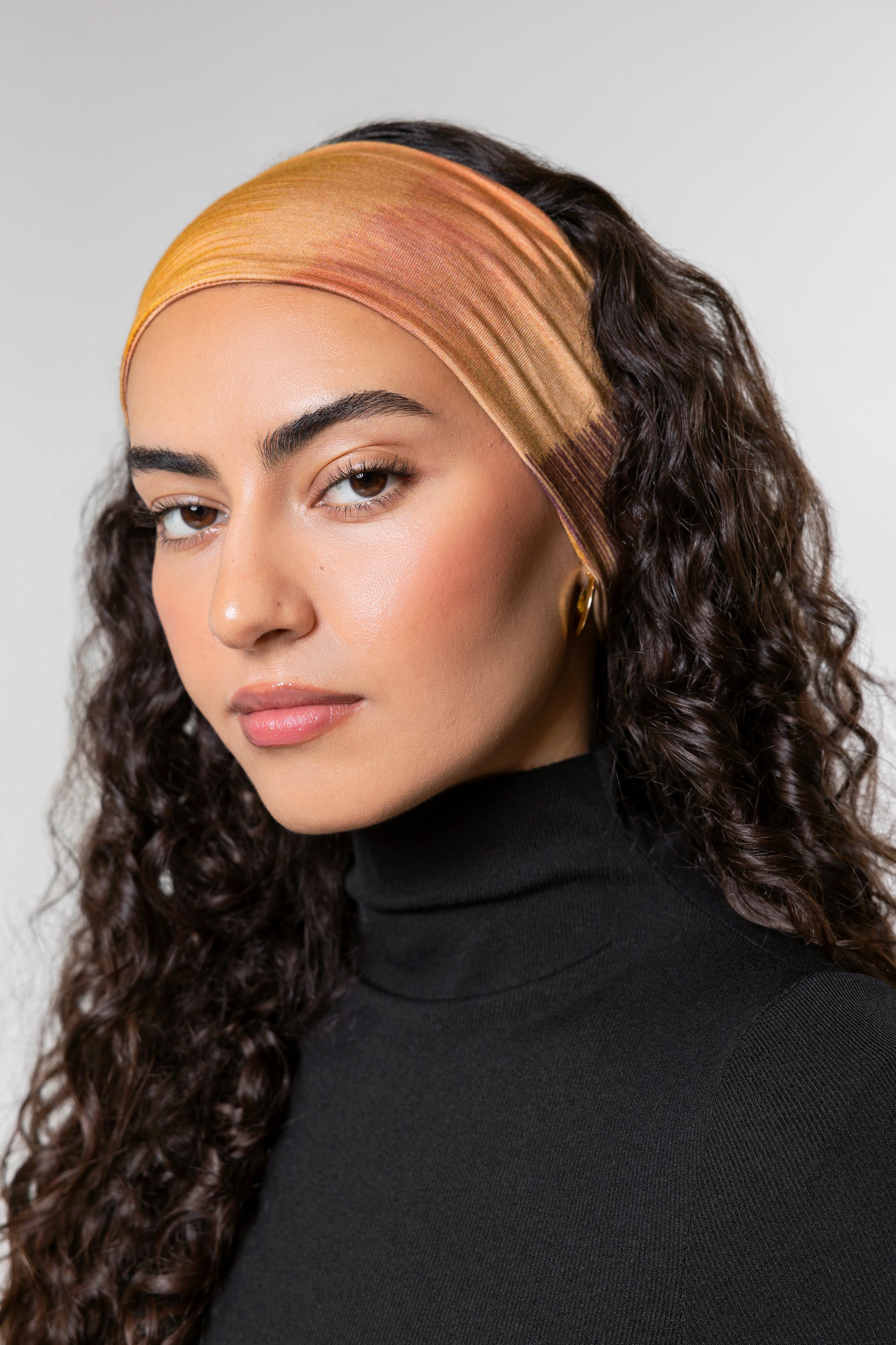 A woman with long, curly dark brown hair wearing a wide, soft fabric headband in warm gradient tones of orange, yellow, and peach, paired with a black turtleneck top against a light gray studio background.