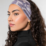 A woman with long, curly dark brown hair is shown in profile wearing a wide, soft fabric headband with a light lavender and gray floral pattern. She is wearing a black turtleneck top and small gold hoop earrings against a neutral light gray background.