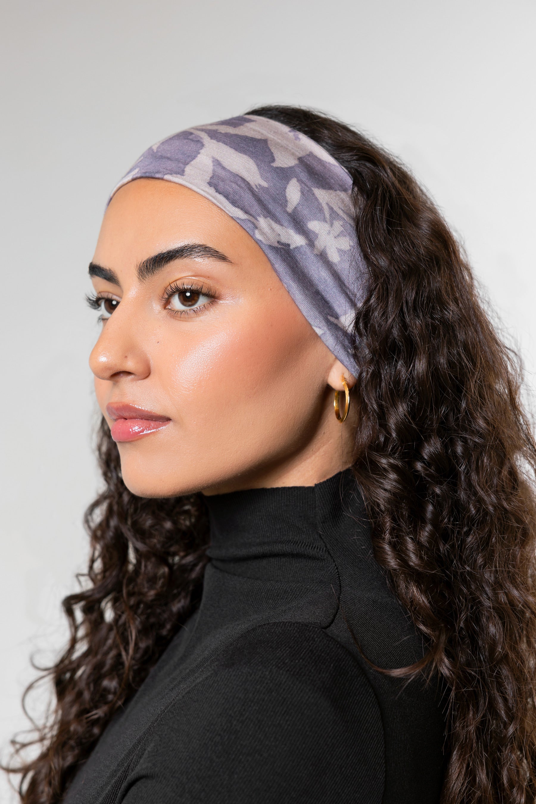 A woman with long, curly dark brown hair is shown in profile wearing a wide, soft fabric headband with a light lavender and gray floral pattern. She is wearing a black turtleneck top and small gold hoop earrings against a neutral light gray background.