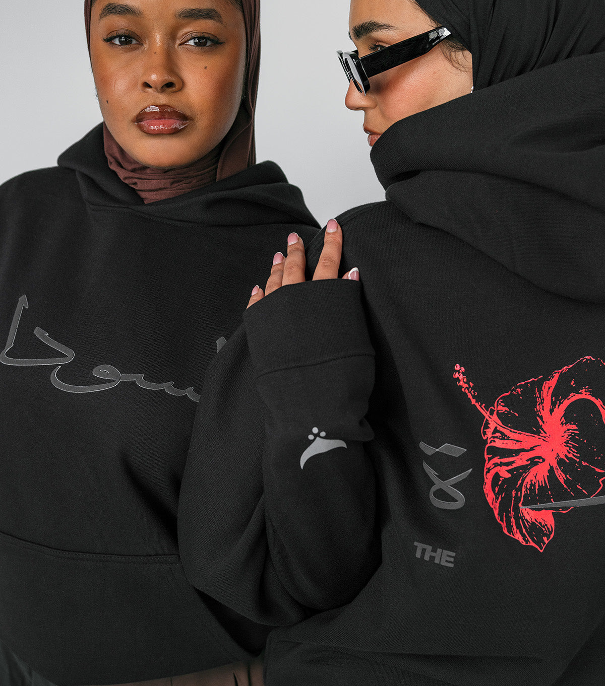 Two people wearing black hoodies with designs on a plain background