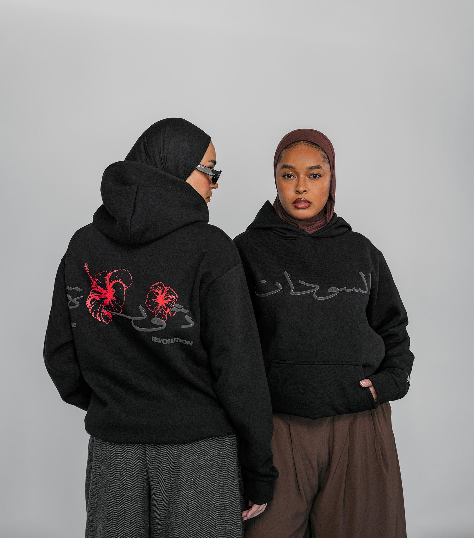 Two women wearing black hoodies with Arabic text and red designs on a gray background