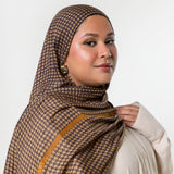A woman wearing a brown and beige houndstooth patterned hijab with a mustard yellow border, paired with a cream-colored top, photographed against a solid light gray background.
