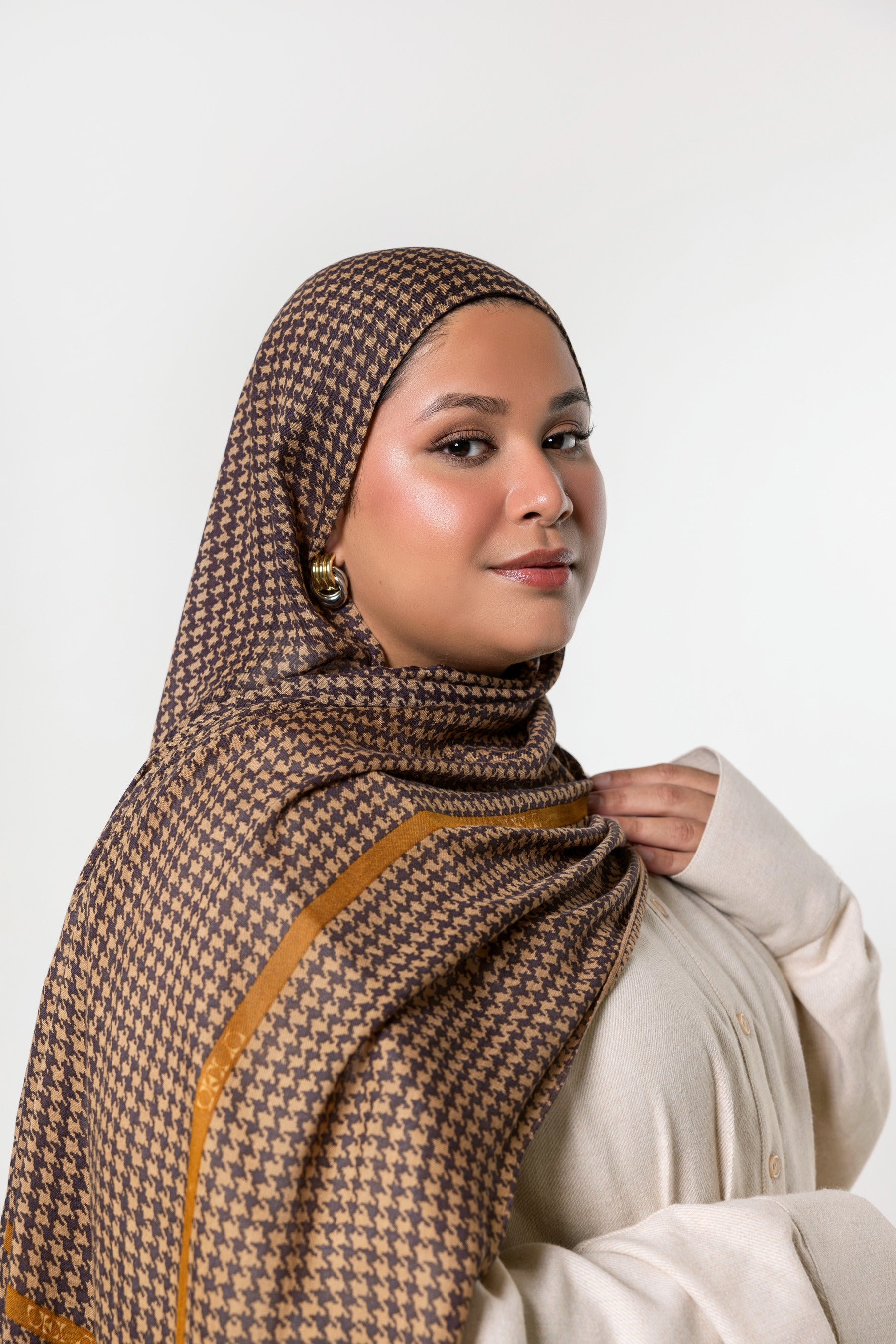 A woman wearing a brown and beige houndstooth patterned hijab with a mustard yellow border, paired with a cream-colored top, photographed against a solid light gray background.