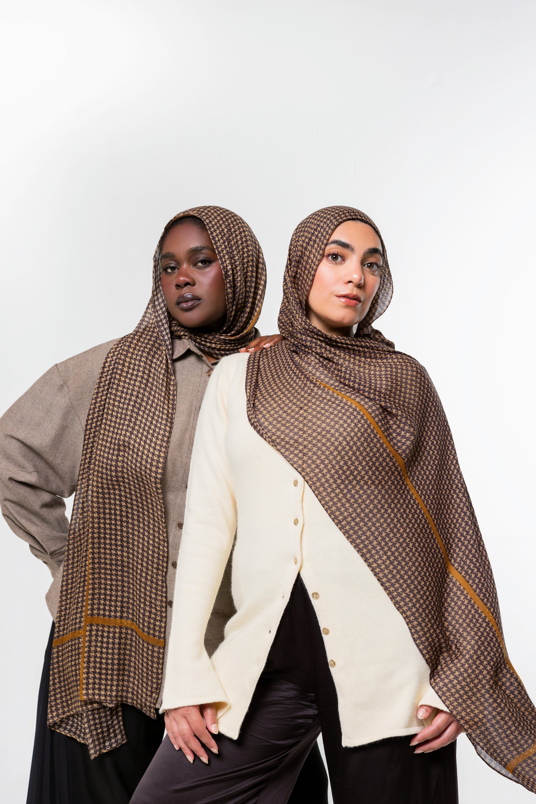 Two models wearing brown patterned hijabs and shawls with gold stripes, styled with neutral-colored tops against a plain white studio background.