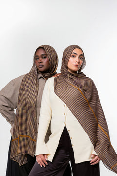 Two models wearing brown patterned hijabs and shawls with gold stripes, styled with neutral-colored tops against a plain white studio background.