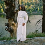 Woman in a white dress standing next to a large tree by a lake with a forest in the background