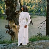 Woman in a white dress standing next to a large tree by a lake with a forest in the background