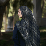 A woman in profile wearing a navy blue dress and a dark gray and navy plaid hijab, standing outdoors in a park with trees and columns in the background.