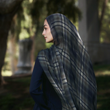 A woman in profile wearing a navy blue dress and a dark gray and navy plaid hijab, standing outdoors in a park with trees and columns in the background.
