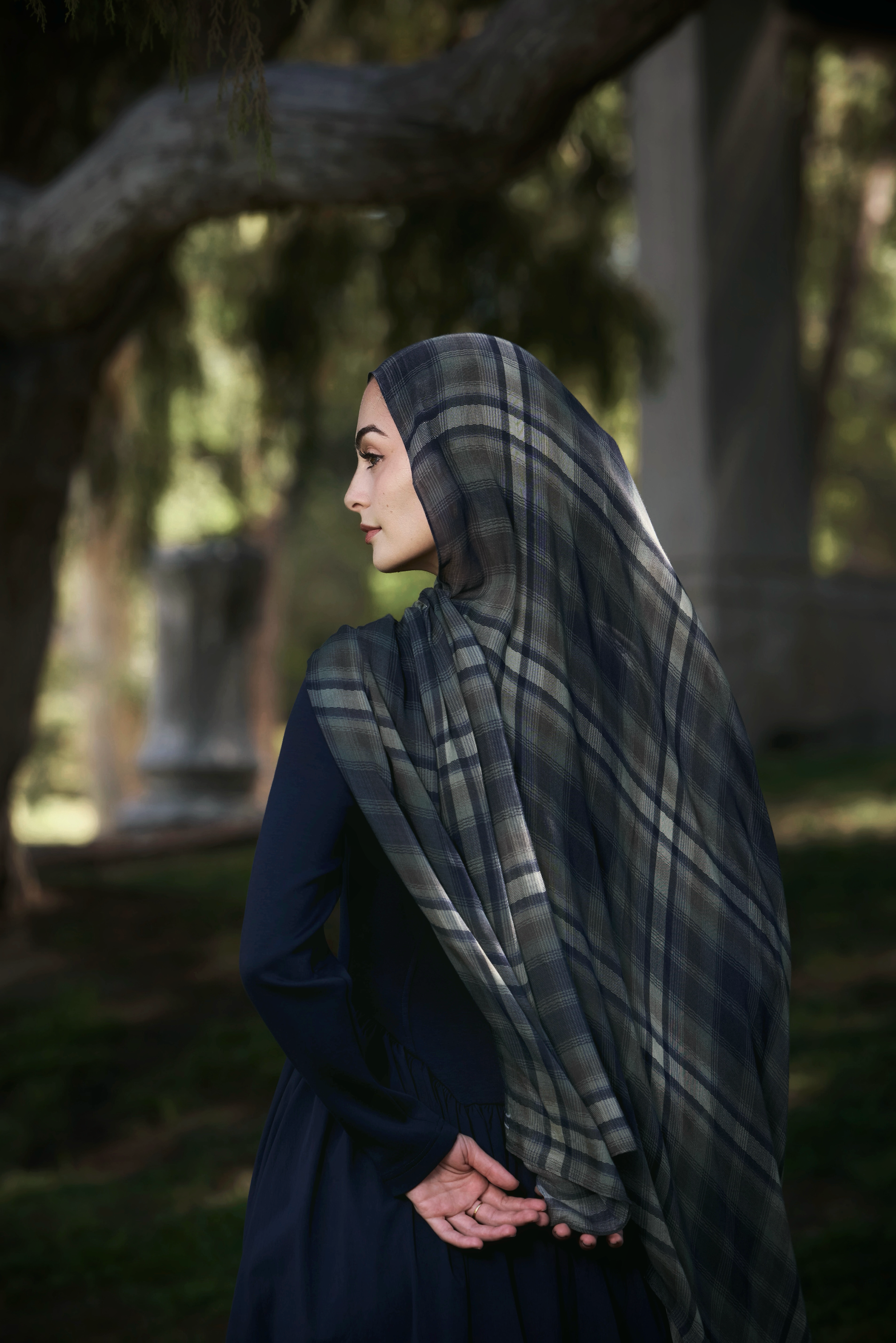 A woman in profile wearing a navy blue dress and a dark gray and navy plaid hijab, standing outdoors in a park with trees and columns in the background.