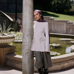 A woman wearing a light pink long-sleeve tunic with lace-up detailing, gray wide-leg pants, black boots, and a purple hijab stands outdoors next to a stone column in front of a garden pond.