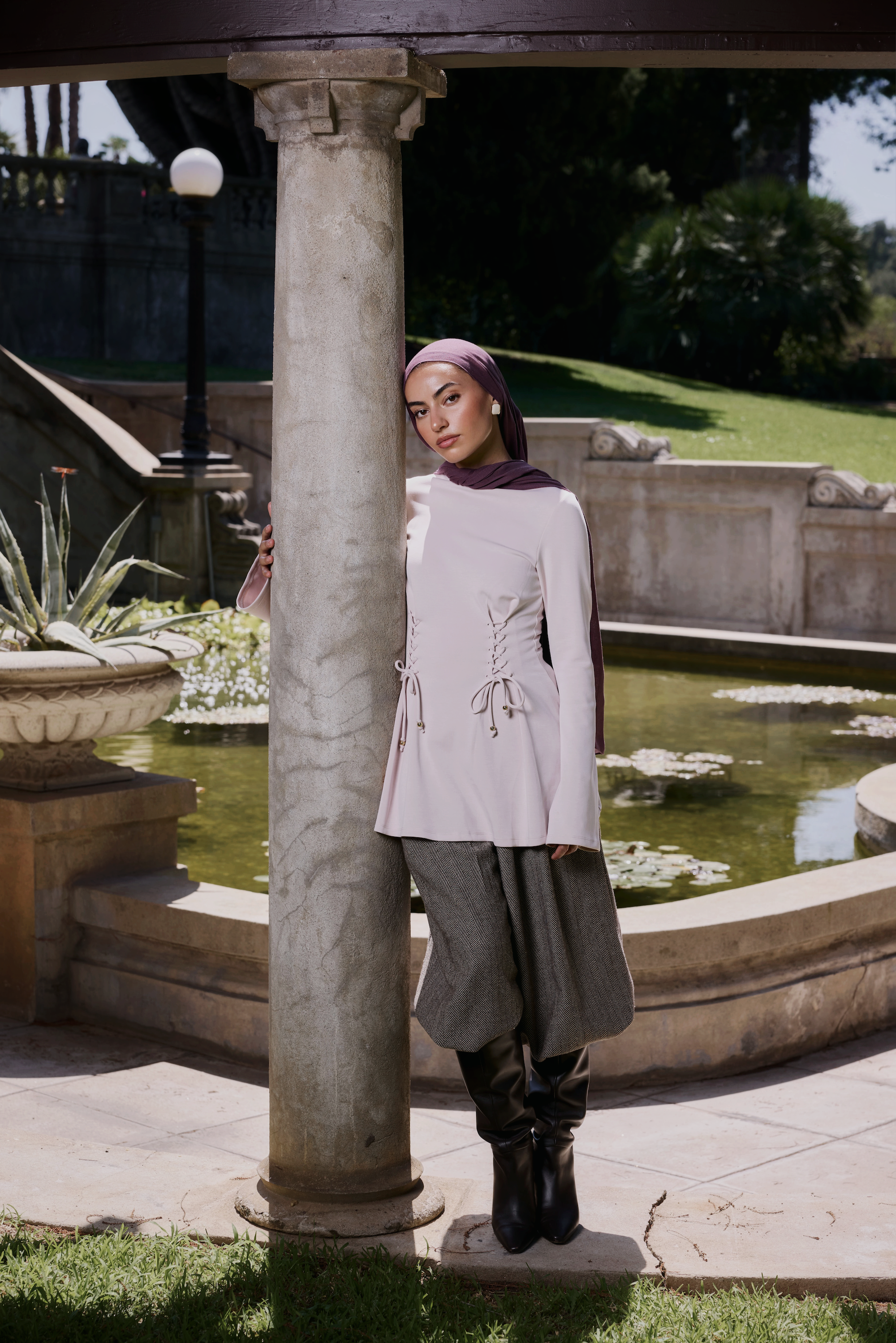 A woman wearing a light pink long-sleeve tunic with lace-up detailing, gray wide-leg pants, black boots, and a purple hijab stands outdoors next to a stone column in front of a garden pond.