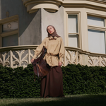 A woman wearing a beige long-sleeve top with a drawstring waist, a dark brown maxi skirt, and a brown headscarf stands outdoors in front of a large beige building with ornate windows and a green hedge.