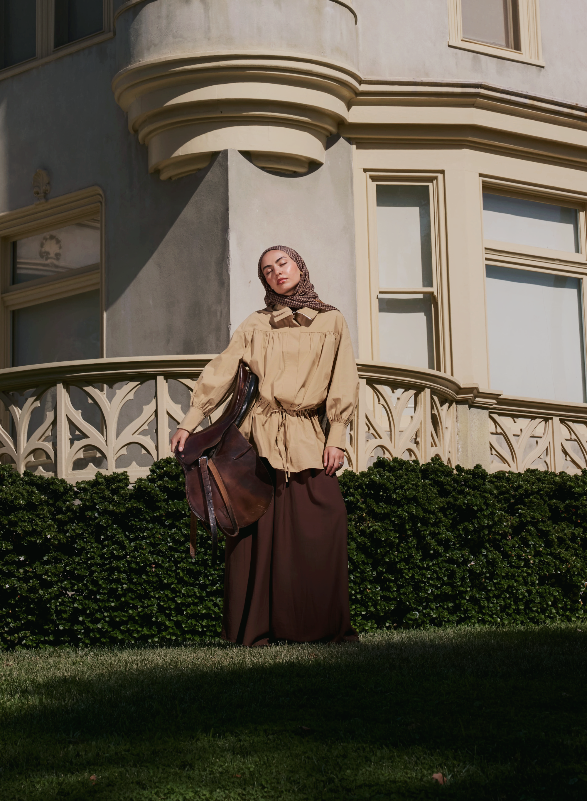 A woman wearing a beige long-sleeve top with a drawstring waist, a dark brown maxi skirt, and a brown headscarf stands outdoors in front of a large beige building with ornate windows and a green hedge.