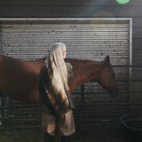 A person in a long white head covering and beige outfit stands beside a brown horse in front of a rustic metal barn door, with a stable interior and hay visible on the ground.