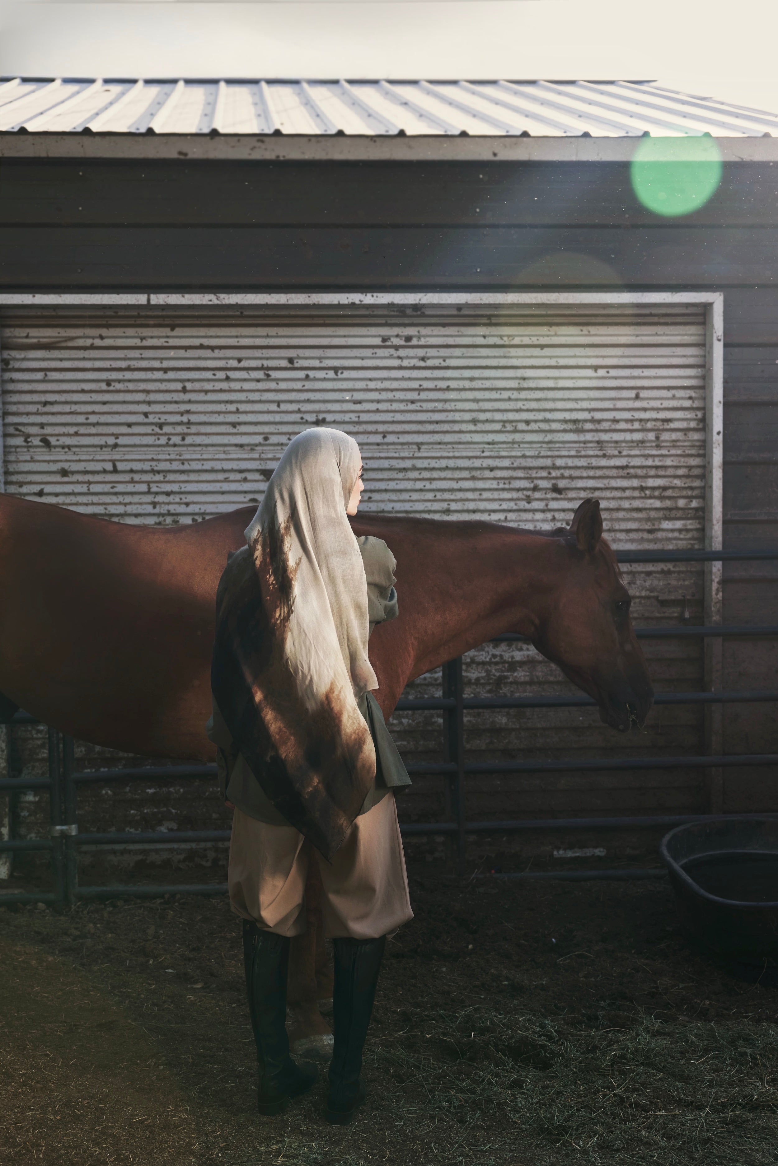 A person in a long white head covering and beige outfit stands beside a brown horse in front of a rustic metal barn door, with a stable interior and hay visible on the ground.
