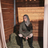 A woman wearing a vintage-style olive green jacket, brown trousers, and a beige hijab poses inside a rustic wooden barn with hay on the floor.