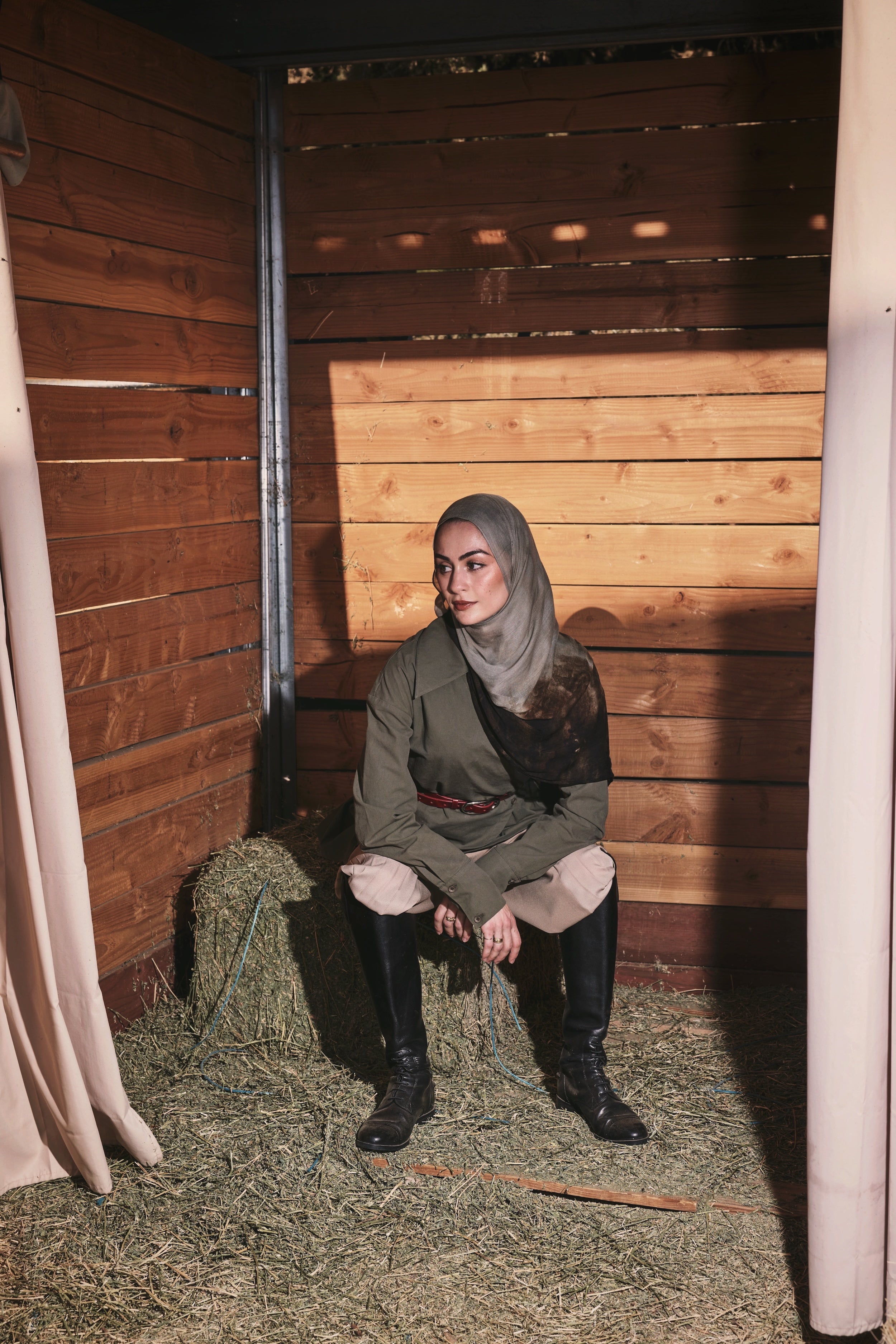 A woman wearing a vintage-style olive green jacket, brown trousers, and a beige hijab poses inside a rustic wooden barn with hay on the floor.