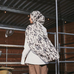 A person standing in profile in a barn-like setting, wearing a white and black patterned hijab, a white long-sleeve top, and loose brown pants.