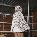 A person standing in profile in a barn-like setting, wearing a white and black patterned hijab, a white long-sleeve top, and loose brown pants.