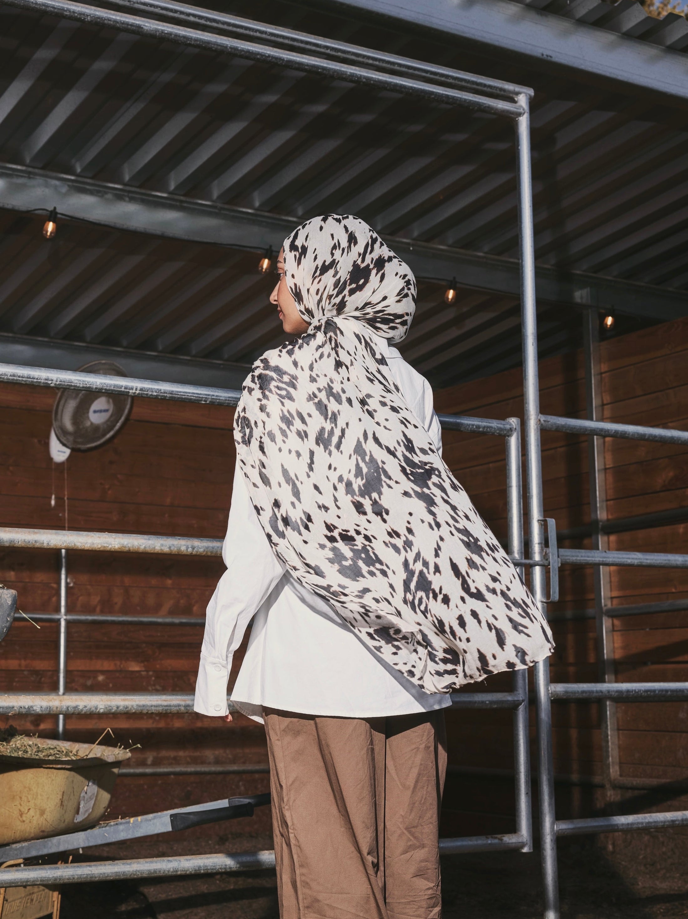A person standing in profile in a barn-like setting, wearing a white and black patterned hijab, a white long-sleeve top, and loose brown pants.