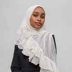 A woman wearing a white ruffled hijab and a black long-sleeve top, photographed against a plain light gray studio background.