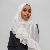 A woman wearing a white ruffled hijab and a black long-sleeve top, photographed against a plain light gray studio background.
