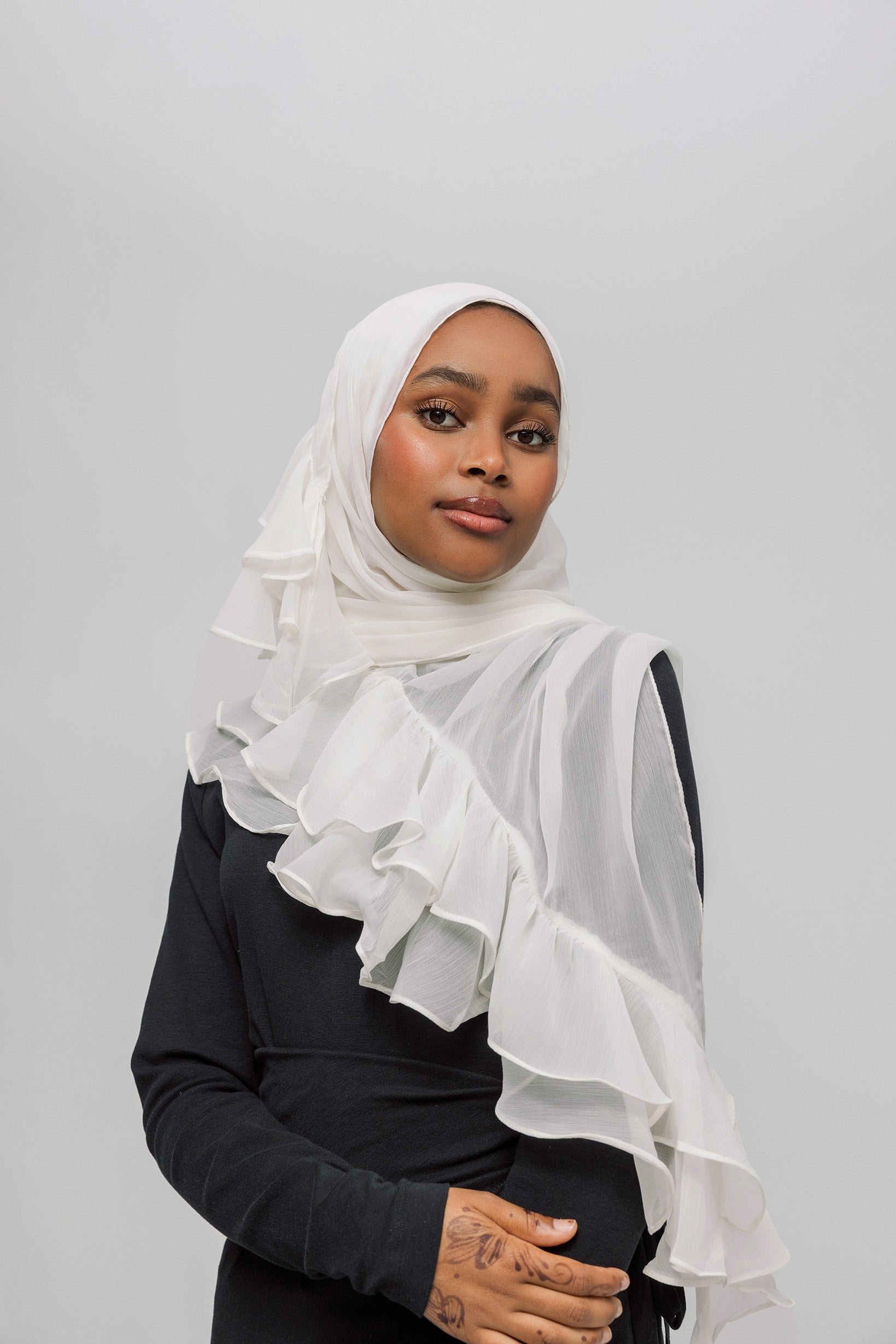 A woman wearing a white ruffled hijab and a black long-sleeve top, photographed against a plain light gray studio background.