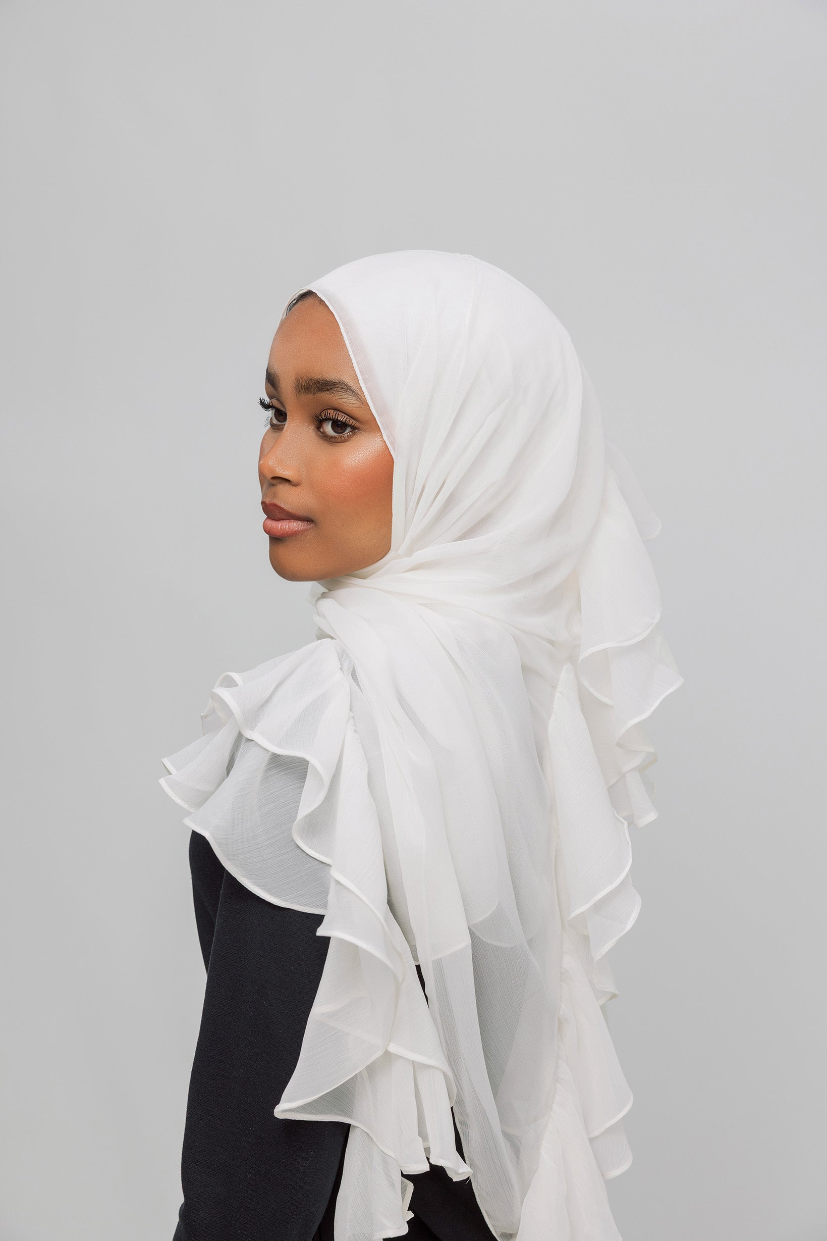 A woman wearing a white ruffled hijab, shown in profile against a plain light gray studio background.