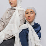 Two women wearing hijabs and long-sleeve satin tops, posed against a light gray studio background. The woman on the left wears a light beige top and a white hijab with layered ruffles; the woman on the right wears a navy blue top and a white hijab with ruffled detailing.