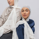Two women wearing hijabs and long-sleeve satin tops, posed against a light gray studio background. The woman on the left wears a light beige top and a white hijab with layered ruffles; the woman on the right wears a navy blue top and a white hijab with ruffled detailing.
