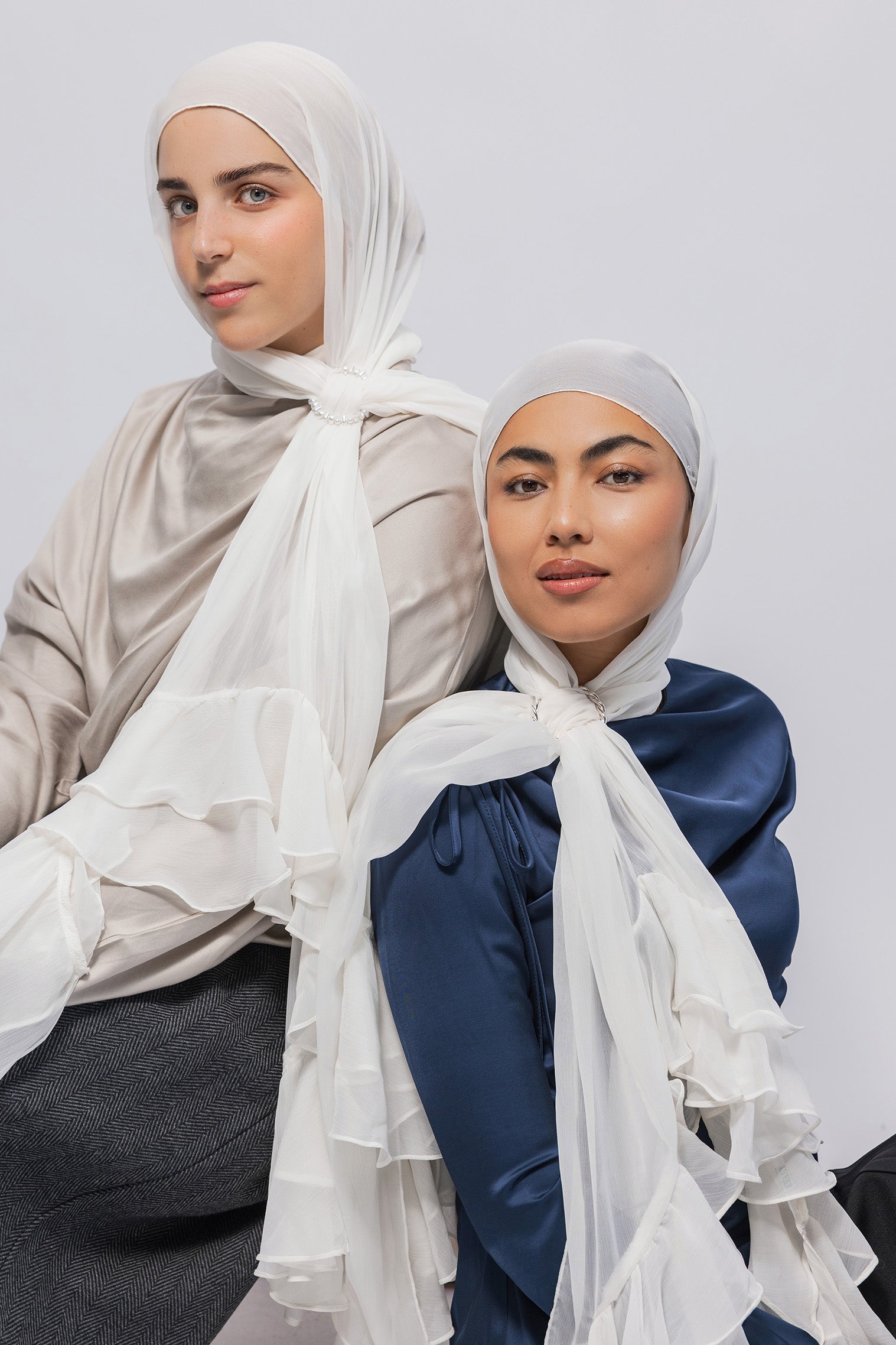 Two women wearing hijabs and long-sleeve satin tops, posed against a light gray studio background. The woman on the left wears a light beige top and a white hijab with layered ruffles; the woman on the right wears a navy blue top and a white hijab with ruffled detailing.