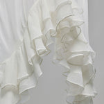 A close-up of a white ruffled fabric, likely a dress or curtain, featuring multiple layers of soft, cascading ruffles against a light gray background.