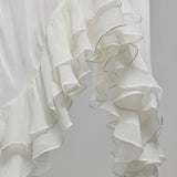 A close-up of a white ruffled fabric, likely a dress or curtain, featuring multiple layers of soft, cascading ruffles against a light gray background.
