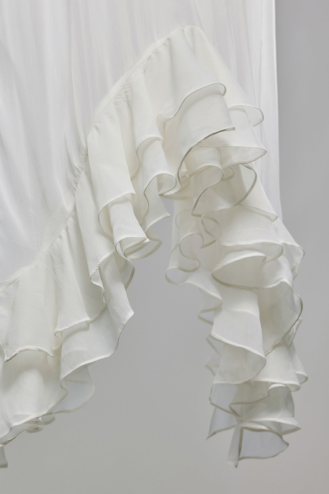 A close-up of a white ruffled fabric, likely a dress or curtain, featuring multiple layers of soft, cascading ruffles against a light gray background.