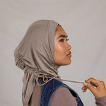 Woman adjusting a taupe grey ruching hijab against a plain background.