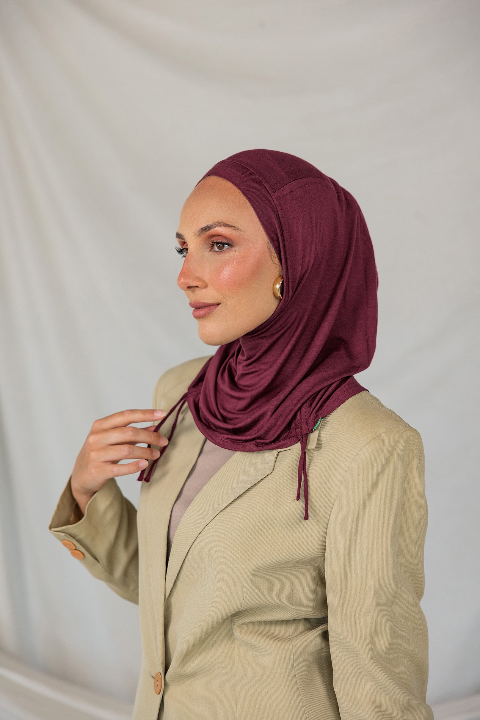 Woman wearing a burgundy ruched hijab and beige blazer against a plain background.