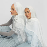 Two women seated side by side wearing white lace-trimmed hijabs and matching pale blue outfits against a plain light gray studio background.