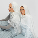 Two women seated side by side wearing white lace-trimmed hijabs and matching pale blue outfits against a plain light gray studio background.