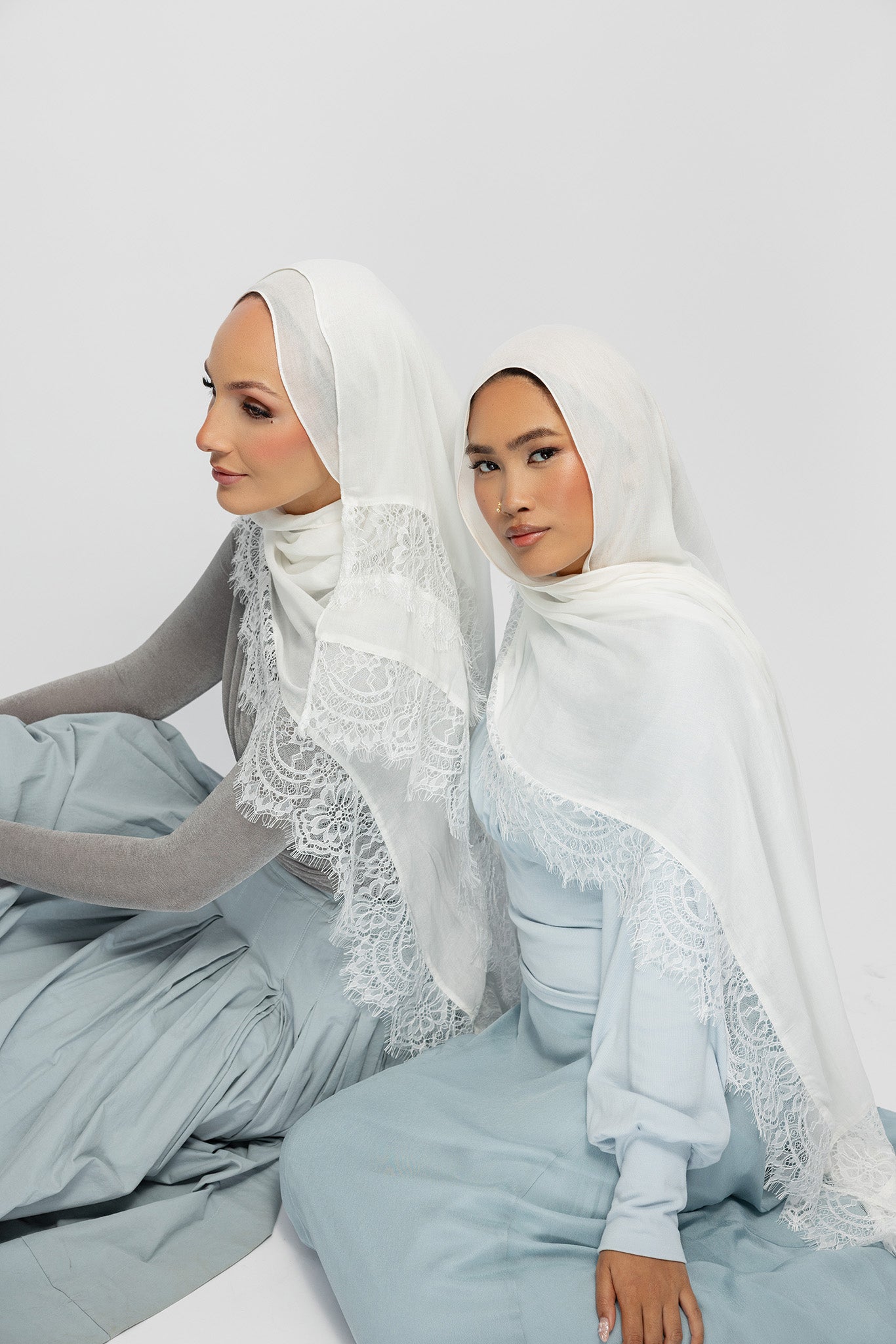 Two women seated side by side wearing white lace-trimmed hijabs and matching pale blue outfits against a plain light gray studio background.