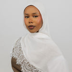 A woman wearing a white hijab with delicate lace trim draped over her shoulder, paired with a brown top, set against a plain light gray background.