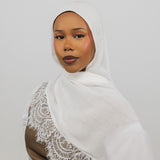 A woman wearing a white hijab with delicate lace trim draped over her shoulder, paired with a brown top, set against a plain light gray background.