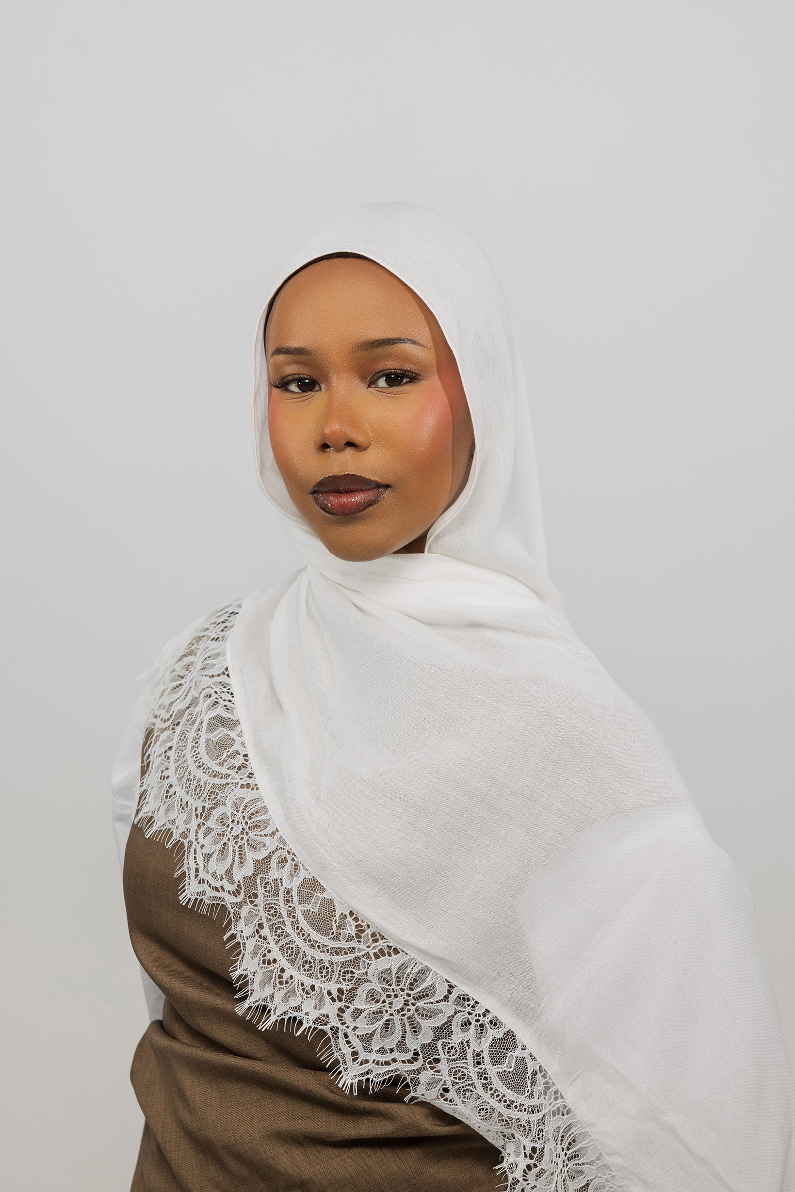 A woman wearing a white hijab with delicate lace trim draped over her shoulder, paired with a brown top, set against a plain light gray background.
