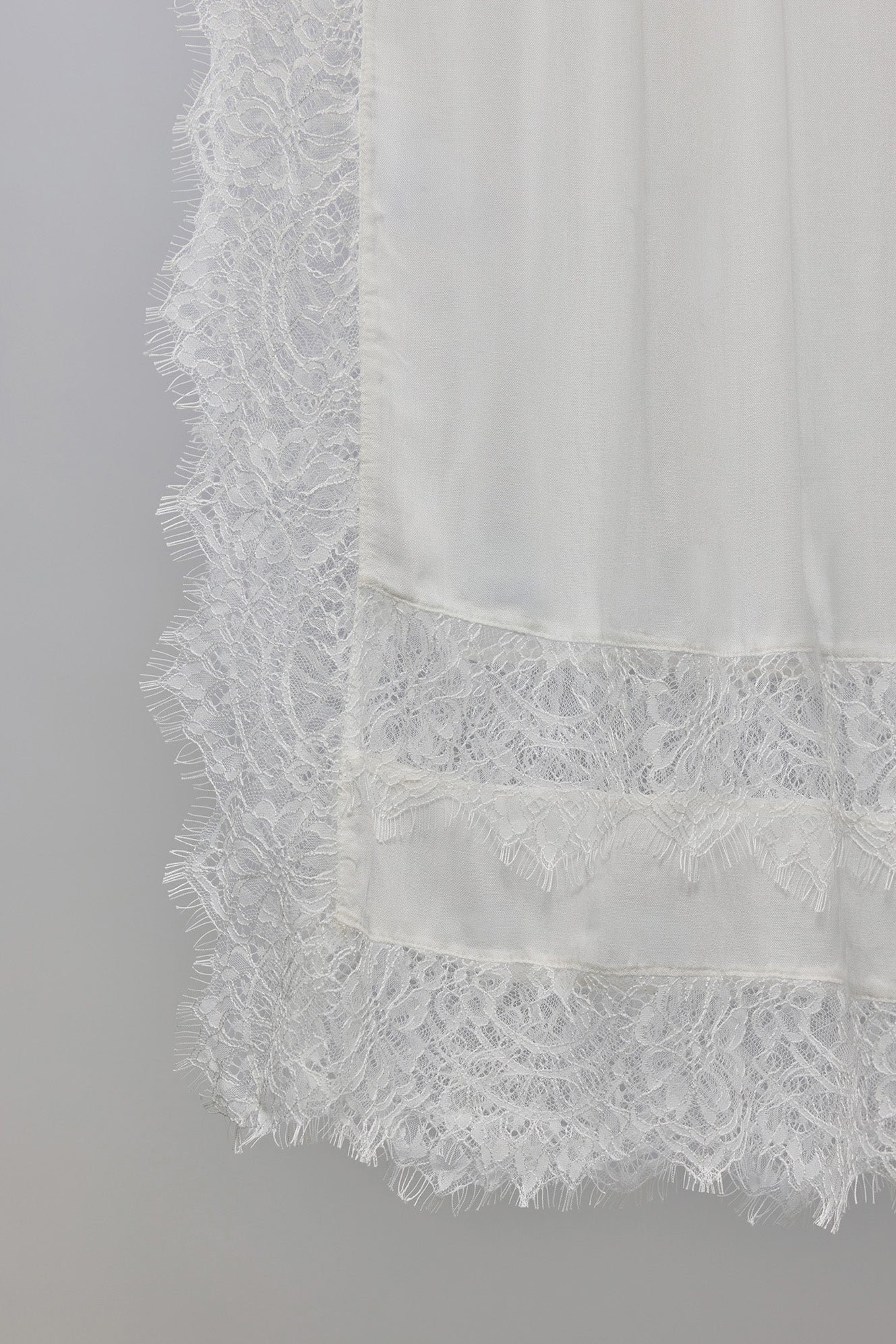 Close-up of a white sheer fabric with a wide lace border and a horizontal band of floral lace near the bottom, shown against a plain light gray background.