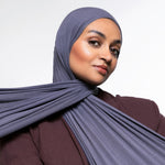 A woman wearing a slate blue-gray hijab and a matching dark plum-brown top, photographed against a clean light gray studio background.